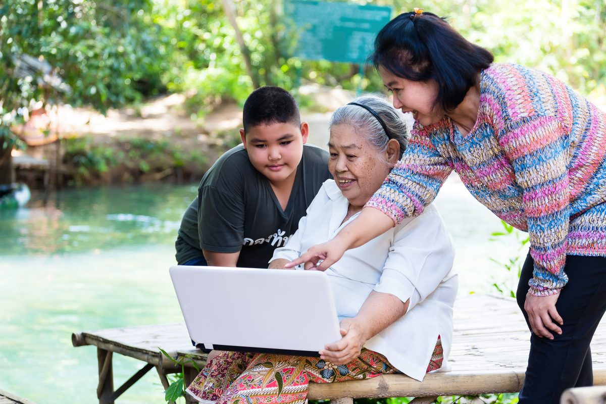 Daughter teaching her mother and little son looking on laptop with cheerful, they are sitting on bamboo couch, near the waterfall  at relaxing time in weekend